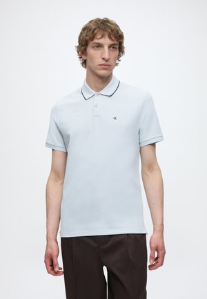 Νέος άνδρας με σγουρά ανοιχτό καφέ μαλλιά που φοράει ένα ανοιχτό μπλε κοντομάνικο πόλο και σκούρο καφέ παντελόνι, στέκεται μπροστά σε απλό φόντο.