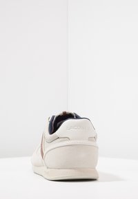 Sneaker bianco in pelle con dettagli marroni, tacco testurizzato e morbida pelle scamosciata. Presenta un logo sul tacco e un interno imbottito.