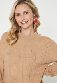 Blondynka uśmiechająca się, ubrana w beżowy sweter o fakturze, duże czerwone kolczyki w kształcie kwiatu i liścia, pozująca na neutralnym tle.