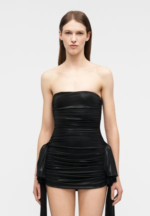 Kobieta o prostych brązowych włosach, ubrana w czarną mini sukienkę bez ramiączek z marszczeniami i bocznymi szarfami, stojąca na tle jasnego, gładkiego tła.