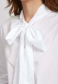 Blusa bianca con dettaglio a nodo al collo, con lunghi nastri fluttuanti e una texture liscia, su uno sfondo chiaro.