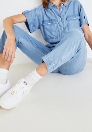 Persoon in een lichtblauwe denim jumpsuit en witte sneakers zit op de vloer met gebogen benen, een arm rustend op de knie tegen een effen achtergrond.