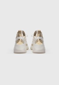 Beige és arany színű sportcipő pár, fehér, vastag talppal, hátulról látható, egyszerű fehér háttéren.