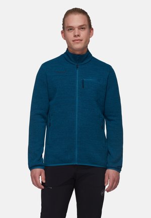 Giovane uomo con capelli chiari che indossa una giacca in pile blu con cerniera e pantaloni neri, in piedi davanti a uno sfondo chiaro semplice.