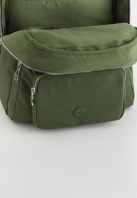 Mochila de nailon verde con un amplio compartimento principal, un bolsillo delantero con cremallera y una insignia de logo circular. Textura suave con un acabado liso.