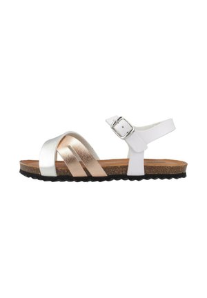 Sandale plate blanche et or rose avec semelle en liège, bride de cheville à boucle et lanières croisées à l'avant sur fond blanc.