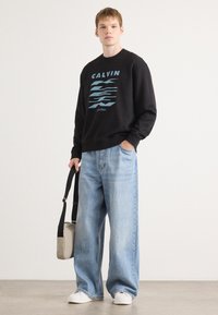 Sweatshirt preto com o logotipo "CALVIN" em azul, combinado com jeans de perna larga azul claro. O modelo segura uma bolsa transversal cinza. Ténis brancos.