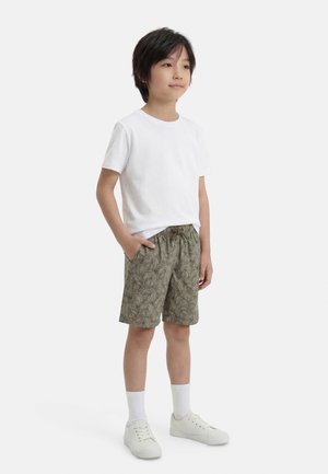 Copil cu păr scurt și închis la culoare, purtând un tricou alb, pantaloni scurți verzi cu model, șosete albe și adidași albi, stând cu mâinile în buzunare.