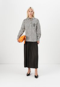 Camicia a righe a maniche lunghe bianca e nera, con una tasca decorata, abbinata a una gonna plissettata nera e una pochette arancione. Tacchi neri.