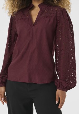 Kobieta ubrana w bordową bluzkę z koronkowymi rękawami typu biskup i dekoltem z rozcięciem, w połączeniu z czarnymi spodniami, widziana od szyi do bioder.