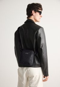 Borsa a tracolla in pelle nera con una texture a motivo e logo Calvin Klein, dotata di tracolla regolabile. Indossata sopra una giacca nera.