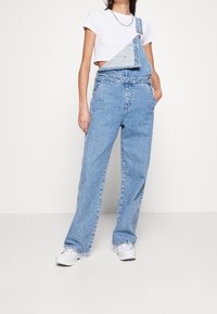 Hellblaue Jeanslatzhose mit einem verkürzten Design, einem verstellbaren Schulterträger, lockeren, weiten Beinen und Seitentaschen.
