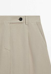 Beige bomuldsbukser har en sort knaplukning, sidelommer og en glat tekstur med subtile stikninger.