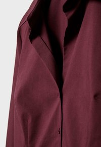 Burgunderfarvet skjorte med en glat tekstur, der har en spids krave, enkelt knaplukning og blødt stof, der hænger langs siden.