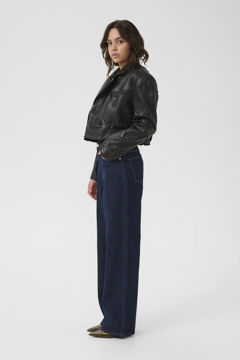 Svart kort läderjacka med en texturerad yta, tillsammans med högmidjade, vida mörk denimjeans och spetsiga gröna skor.