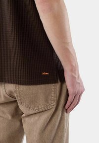 Brun ribbad skjorta med en orange logotyp vid fållen, tillsammans med ljusbeige byxor med bakfickor. Handen vilar på byxans sida.