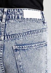 Pantalones vaqueros de mezclilla con un acabado azul desgastado, que cuentan con un parche de cuero blanco en la cintura, bolsillos con costuras múltiples y un corte relajado.