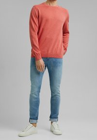 Helles Korallenpullover, Rundhalsausschnitt, lange Ärmel, gerippter Saum. Getragen mit hellblauen Slim-Fit-Jeans und weißen Sneakers. Einfacher, strukturierter Stoff.