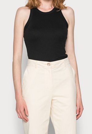 Personne portant un haut noir ajusté sans manches et un pantalon crème taille haute avec des boutons à l'avant et des poches latérales.