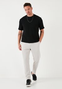 Schwarzes kurzärmliges Shirt mit Struktur, kombiniert mit hellgrauen, taillierten Hosen und schwarzen Sneakern mit Riemendetails, Modell steht vor einem weißen Hintergrund.