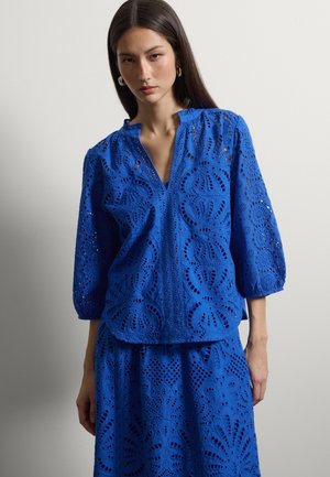 Mujer que lleva una blusa azul de encaje con ojales, mangas abullonadas tres cuartos y falda a juego, de pie frente a un fondo liso gris claro.