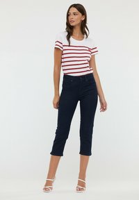 T-shirt blanc à manches courtes avec des rayures horizontales rouges, associé à un pantalon en denim foncé raccourci. Le modèle porte des sandales blanches à lanières.