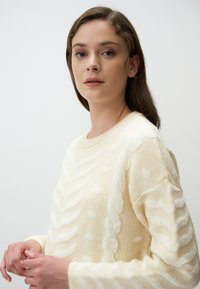 Maglione color crema con motivi bianchi testurizzati, con scollo rotondo e maniche lunghe, realizzato in morbido e leggero materiale lavorato a maglia.