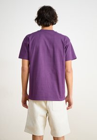 Camiseta de algodón morada de manga corta, con cuello redondo y una textura suave, combinada con pantalones cortos de color claro que tienen un bolsillo en la parte trasera.