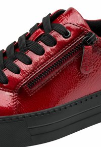 Red patent leather sneaker with a textured surface, black laces, side zipper, and a chunky black rubber sole.