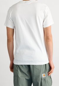 Camiseta de algodón blanca con un escote redondo clásico, corte relajado y mangas cortas, combinada con pantalones cargo verdes que presentan un parche de logo.