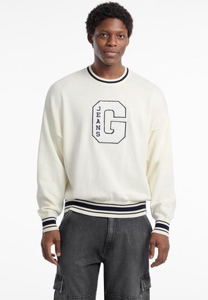 Cremfarbener Pullover mit dunkelblauen Streifen am Kragen und an den Bündchen, mit einem großen gestickten "G" und "JEANS" darunter. Weicher Stoff, lockere Passform.