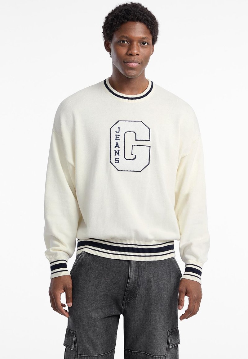 Suéter crema con rayas marinas en el cuello y los puños, que presenta una gran "G" bordada con "JEANS" debajo. Textura suave, corte relajado.