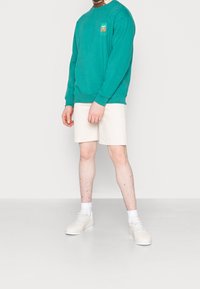 Sweatshirt turquoise avec un petit logo, assorti à un short beige. Des baskets blanches et des chaussettes mi-mollet complètent la tenue. Design simple et décontracté.