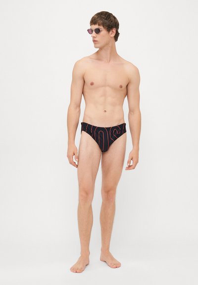 Jeune homme portant un maillot de bain noir avec un texte en rouge, debout pieds nus contre un fond blanc uni, portant des lunettes de soleil.