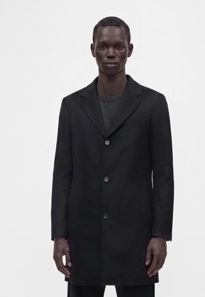 Homme portant un manteau noir à simple boutonnage sur une chemise gris foncé, debout devant un fond blanc uni.