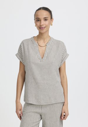 Žena oblečená v béžovom tričku s krátkymi rukávmi a zodpovedajúcich nohaviciach s tenkými zvislými čiernymi pruhmi, so zlatými kruhovými náušnicami a zlatým náhrdelníkom, stojaca a usmievajúca sa.