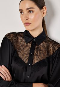 Maglietta nera con yoke in pizzo, bottone frontale e finitura in satin liscia. I dettagli notevoli includono bordi in pizzo e bottoni neri.