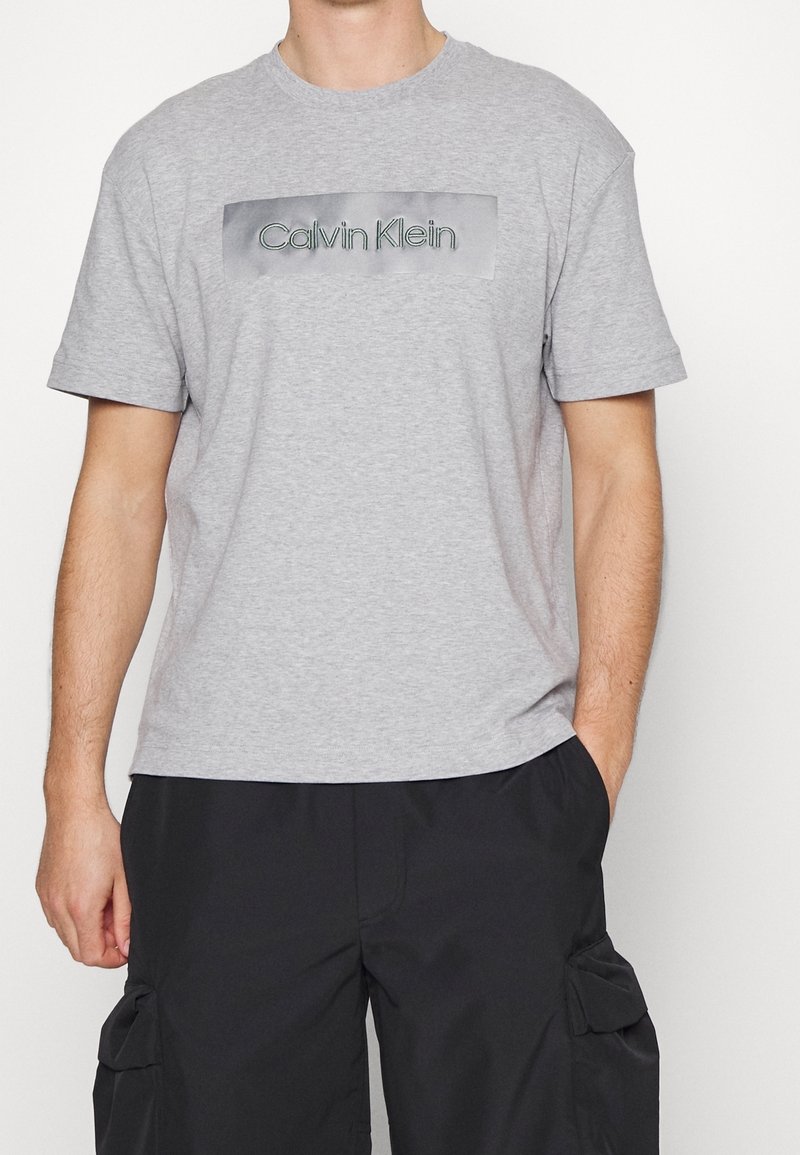 Graues Baumwoll-T-Shirt mit kurzen Ärmeln und einem zentrierten rechteckigen Motiv, auf dem "Calvin Klein" in Schwarz auf grauem Hintergrund steht.