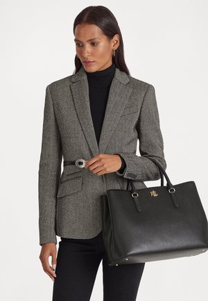 Femme ajustant une ceinture noire portée sur un blazer à chevrons gris, portant un grand sac cabas en cuir noir avec un logo RL doré.