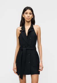 Γυναίκα που φοράει μαύρο, διπλής σειράς, μίνι φόρεμα σακάκι χωρίς μανίκια με γιακά, ζώνη και μπροστινές τσέπες, στέκεται μπροστά σε απλό φόντο.