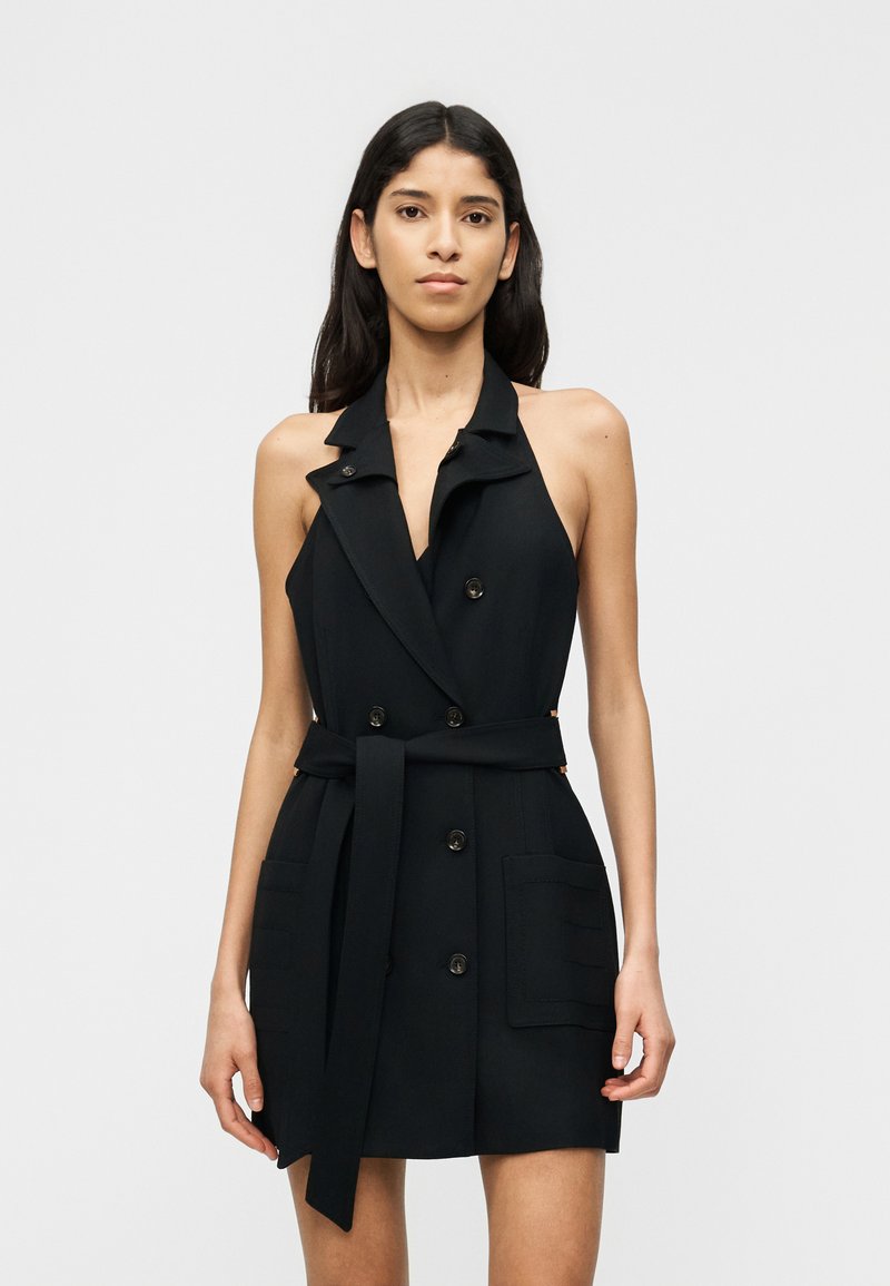 Γυναίκα που φοράει μαύρο, διπλής σειράς, μίνι φόρεμα σακάκι χωρίς μανίκια με γιακά, ζώνη και μπροστινές τσέπες, στέκεται μπροστά σε απλό φόντο.