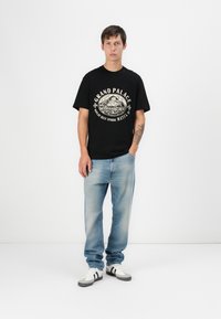 T-shirt nera in cotone con scollo rotondo e stampa grafica color crema; jeans in denim azzurri; sneakers bianche con strisce nere.