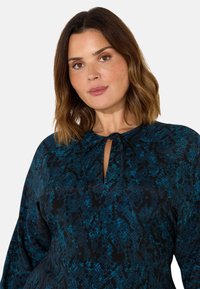 Blus med blå och svart mönster, knytkrage, långa ärmar och en texturerad tyg. Har en nyckelhålöppning vid halsringningen.