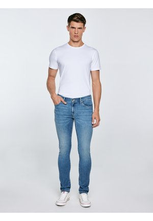 Noor mees, kes seisab ühe käega taskus, kandes valget t-särki, siniseid kitsaid teksaseid ja valgeid tossud, ühevärvilise tausta ees.