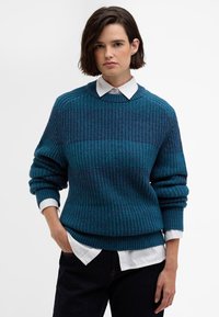 Τυρκουάζ πλεκτό πουλόβερ με υφή, με ραμμένα μανίκια και τελειώματα, φορεμένο πάνω από ένα λευκό πουκάμισο με γιακά. Χαλαρή, casual εφαρμογή.