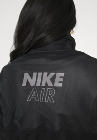 Kvinna med långt mörkt hår som bär en svart mesh Nike Air-jacka, sedd bakifrån, med guld triangulära örhängen synliga.