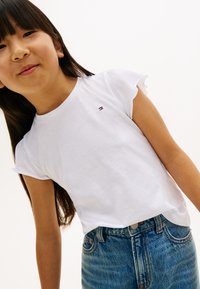 Weißes kurzärmeliges T-Shirt mit Rüschenärmeln und einem kleinen Logo; kombiniert mit blauen Denim-Jeans. T-Shirt aus glattem Stoff.