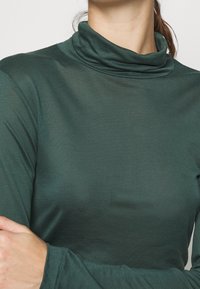 Maglia a maniche lunghe a collo alto di colore verde scuro, caratterizzata da una texture morbida e liscia e una vestibilità rilassata attorno al collo e alle spalle.