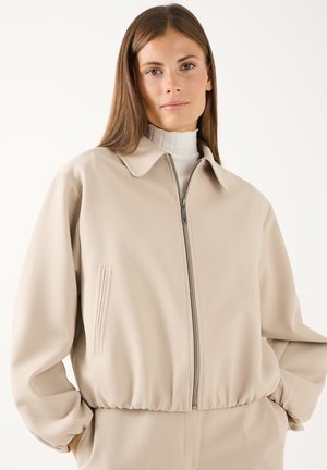 Frau trägt eine beige Jacke mit Reißverschluss über einem weißen Rollkragenpullover, steht mit den Händen in den Taschen vor einem einfarbigen hellen Hintergrund.