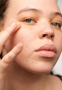 Klart gelé droppar applicerade under ögat, med ljus hudton, fräknar och en septum-piercing. Närbild av ansiktsdrag och hand.